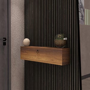 Puede incluir: Una estantería flotante de madera con un acabado cálido y natural está montada en una pared oscura con listones verticales. Un pequeño objeto decorativo redondo de color crema se encuentra a la izquierda, y un terrario de vidrio con plantas a la derecha.