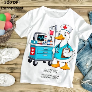Puede incluir: Camiseta blanca con un pato de dibujos animados vestido de enfermero, con una bata azul y un gorro con una cruz roja. El pato está junto a un carrito médico con un monitor y un goteo intravenoso. El texto "BOUT TO CRASH OUT" está debajo.