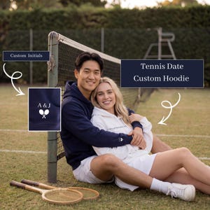 May include: A couple wearing custom hoodies on a tennis court. The navy blue hoodie has the initials "A&J" with crossed tennis rackets. The white hoodie has text on the sleeve. Two vintage tennis rackets rest on the grass.