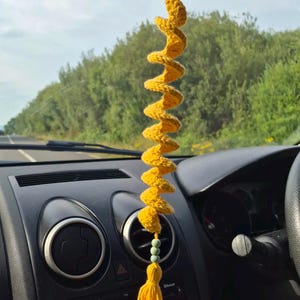 May include: A yellow crocheted spiral hanging ornament suspended from a rearview mirror. The spiral features a textured yarn construction, accented with three small light green beads and a long yellow tassel at the bottom end.
