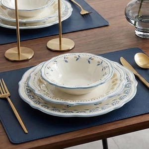 Puede incluir: Una mesa de comedor con platos estampados florales, cubiertos dorados y manteles individuales azules. Dos candelabros dorados con velas blancas están colocados sobre la mesa. Un jarrón de cristal con flores completa la elegante decoración.