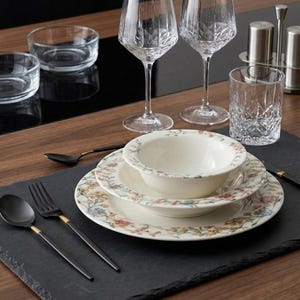 May include: A table setting featuring floral patterned plates, bowls, and black flatware with gold accents. Crystal wine glasses, a tumbler, and small glass bowls are also present. The setting is on a black slate placemat.