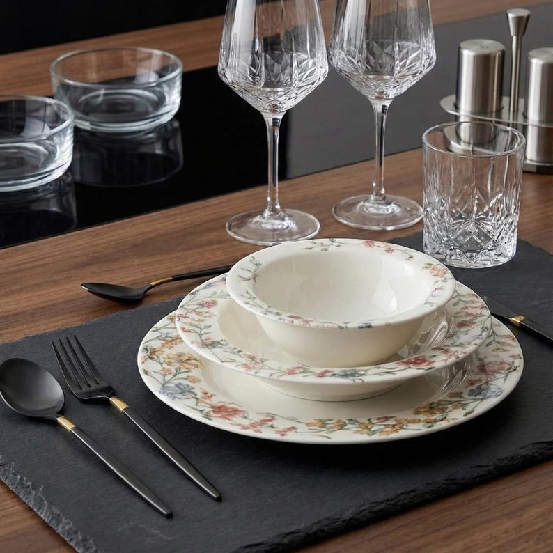 May include: A table setting featuring floral patterned plates, bowls, and black flatware with gold accents. Crystal wine glasses, a tumbler, and small glass bowls are also present. The setting is on a black slate placemat.