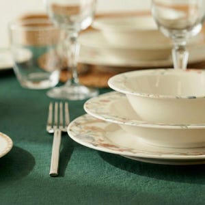 May include: A dining table setting with white ceramic plates and bowls featuring a floral design. The plates and bowls are stacked, with a fork, wine glasses, and a water glass on a green tablecloth.