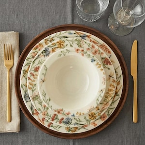 May include: A table setting with a wooden charger, floral-patterned plates, a bowl, and gold cutlery. The plates feature a delicate design of colorful flowers and greenery. A folded linen napkin and glassware complete the elegant arrangement.