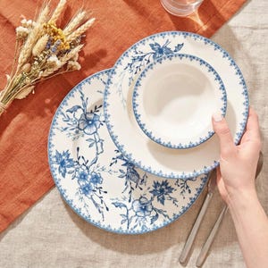May include: A set of white ceramic dinnerware with a blue floral design. The set includes a large plate, a smaller plate, and a bowl. The plates and bowl have a blue border. A hand is holding the bowl.