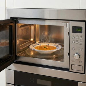 May include: A stainless steel microwave oven with the door open, revealing a bowl of soup on a plate. The plate is on a glass turntable. Steam rises from the soup, indicating it is hot. The microwave's control panel is visible.