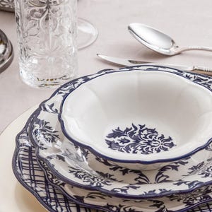 Puede incluir: Un juego de cuenco y platos de cerámica blanca con un patrón floral azul oscuro y bordes festoneados. El cuenco está apilado sobre dos platos, con el plato inferior mostrando un patrón de cuadrícula azul. El set incluye un vaso y cubiertos.