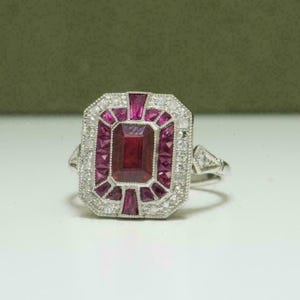 May include: A silver ring featuring a large, rectangular ruby center stone surrounded by smaller rubies and clear accent stones. The ring has a geometric design with a triangular accent on the side.