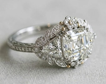 Könnte beinhalten: Ein silberfarbener Ring mit einem großen quadratischen Mittelstein, der von zwei dreieckigen Seitensteinen flankiert wird. Die Ringschiene und die Fassung sind mit kleinen Pavé-Steinen in Blatt- und Blumenmustern sowie goldenen Akzenten verziert.