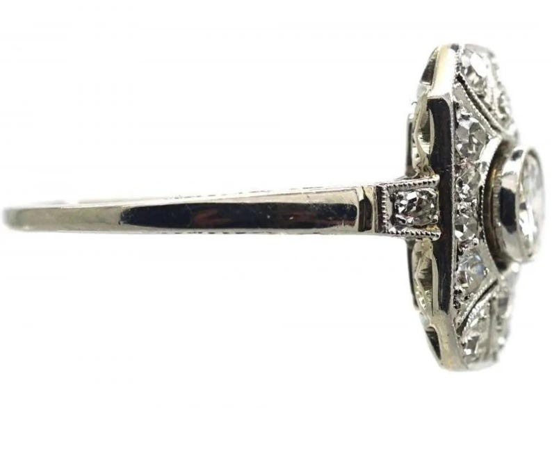 May include: A vintage-style ring with a silver band and a rectangular face. The face is adorned with multiple small diamonds and a larger, round diamond. The ring is set against a white background.