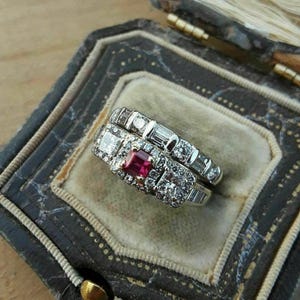 May include: Two silver rings with diamonds and a ruby center stone, displayed in a vintage jewelry box. The rings feature a combination of square and baguette-cut diamonds. The box is dark with a gold clasp.