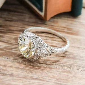 May include: A vintage-style engagement ring with a large, round, light yellow diamond center stone. The ring features intricate filigree and small accent diamonds set in a white metal band, likely platinum or white gold.
