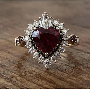 Può includere: Un anello d'oro con una gemma rosso scuro a forma di cuore al centro, circondata da gemme bianche e nere. Il design dell'anello include ulteriori gemme rosse sui lati.