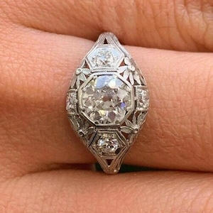 May include: An ornate, vintage-style ring featuring a large, round, clear diamond as the centerpiece. The ring has a detailed, filigree design with smaller diamonds and floral accents, set in a silver or white gold band.