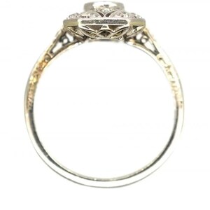 May include: A vintage-style engagement ring with a central diamond and smaller surrounding stones. The ring features an ornate, filigree design on the band and setting, with a silver or white gold finish. The ring's design is symmetrical and detailed.