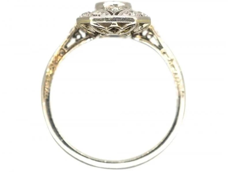 May include: A vintage-style engagement ring with a central diamond and smaller surrounding stones. The ring features an ornate, filigree design on the band and setting, with a silver or white gold finish. The ring's design is symmetrical and detailed.