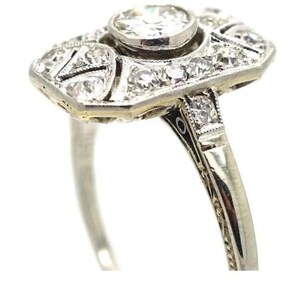 May include: A vintage-style ring with a rectangular face and a round center stone. The ring features numerous small, sparkling diamonds set in a silver or white gold band. The design is intricate and detailed, with a classic, elegant aesthetic.