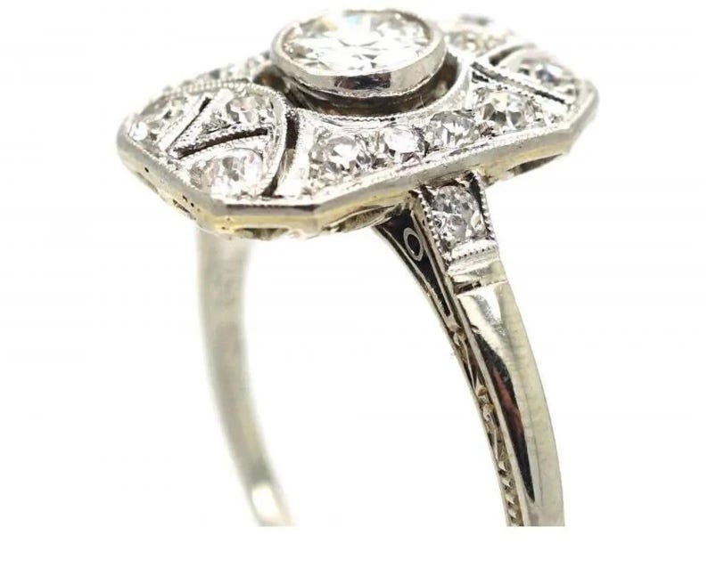 May include: A vintage-style ring with a rectangular face and a round center stone. The ring features numerous small, sparkling diamonds set in a silver or white gold band. The design is intricate and detailed, with a classic, elegant aesthetic.