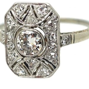 May include: A vintage-style ring with a rectangular face, featuring a large, round, brilliant-cut diamond at the center. The face is surrounded by smaller diamonds in a geometric pattern, set in a white metal band. The ring has a classic, elegant design.