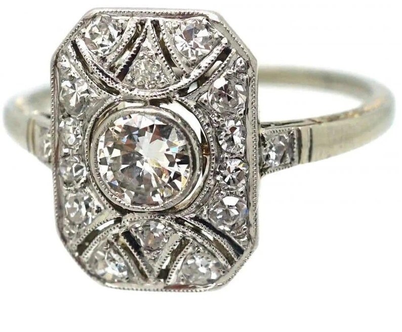May include: A vintage-style ring with a rectangular face, featuring a large, round, brilliant-cut diamond at the center. The face is surrounded by smaller diamonds in a geometric pattern, set in a white metal band. The ring has a classic, elegant design.