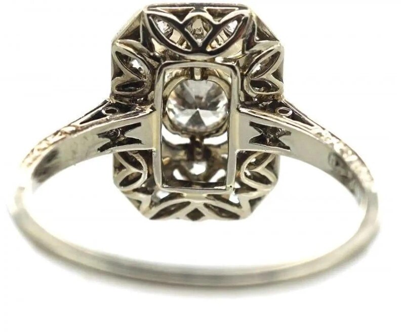 May include: A silver ring with an octagonal setting and a central, round, clear gemstone. The setting features intricate filigree and geometric designs. The band is thin and polished, with decorative elements near the setting.