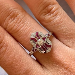 May include: A silver ring with a geometric design featuring red gemstones and small clear stones. The ring has a central round clear stone. The ring is on a finger.