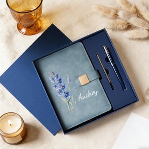 Peut inclure: Un journal bleu-vert avec un motif floral et le nom "Audrey" en lettres blanches, présenté dans un coffret bleu marine. L'ensemble comprend un stylo et un stylet assortis. Une bougie allumée et un verre sont en arrière-plan.
