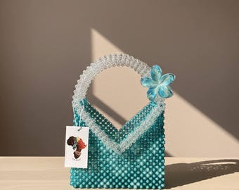 May include: A teal beaded handbag with a clear beaded handle and trim. A light blue flower adorns the side. A tag with an illustration hangs from the handle. Scattered teal and clear beads are on the wooden surface.