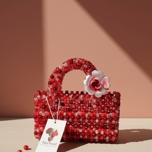 K&ouml;nnte beinhalten: Eine rote, perlenbesetzte Handtasche mit passendem Griff und einer wei&szlig;-rosa Blume. Die Tasche ist rechteckig und tr&auml;gt ein Etikett mit der Aufschrift "Tais Pearl". Lose Perlen liegen am Boden verstreut.