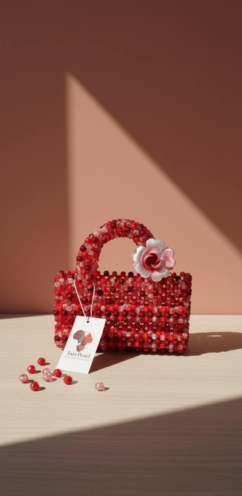K&ouml;nnte beinhalten: Eine rote, perlenbesetzte Handtasche mit passendem Griff und einer wei&szlig;-rosa Blume. Die Tasche ist rechteckig und tr&auml;gt ein Etikett mit der Aufschrift "Tais Pearl". Lose Perlen liegen am Boden verstreut.