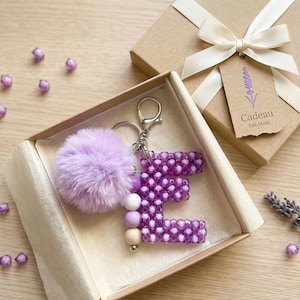 May include: A keychain featuring a letter E made of purple beads, a fluffy light purple pom-pom, and a strand of three smooth beads in shades of purple, white, and beige, all presented in a cardboard gift box with a white ribbon and a tag.