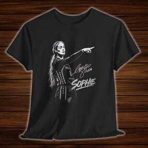 Puede incluir: Una camiseta negra de manga corta con una ilustración gráfica blanca de una jugadora de baloncesto señalando hacia adelante. El diseño incluye una firma y el nombre SOPHIE impreso en una fuente blanca estilizada y negrita en el pecho.