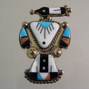 May include: A silver-toned metal pin in the shape of a stylized bird. The pin features inlaid turquoise, black, white, and orange stones. The bird's body is a white triangle with a turquoise accent. The wings have black, orange, and turquoise accents.