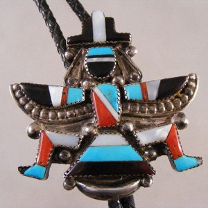 May include: A silver bolo tie featuring a colorful inlaid design. The pendant showcases a stylized bird or figure with turquoise, black, white, and red accents. The bolo tie has a black braided leather cord.