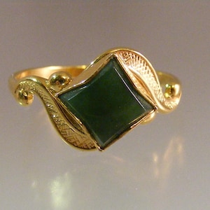 May include: A gold ring with a square, dark green gemstone at the center. The ring has ornate, swirling gold designs on either side of the gemstone. The band is a simple, polished gold.