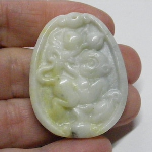 May include: Oval-shaped, off-white pendant with a carved design featuring two figures. The pendant has a light green hue in areas. The figures appear to be in a playful or symbolic pose, set within a detailed border.