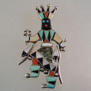 May include: A silver and turquoise Kachina doll pin with a black face, red lips, and a crown. The doll is wearing a turquoise, white, and black outfit with a red and white sash. The pin is made of sterling silver and is inlaid with turquoise, coral, and mother-of-pearl.