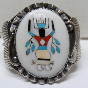 May include: Sterling silver cuff bracelet with an oval, inlaid centerpiece. The centerpiece features a Native American figure with turquoise, red, black, and white details. The bracelet has a detailed, textured design.