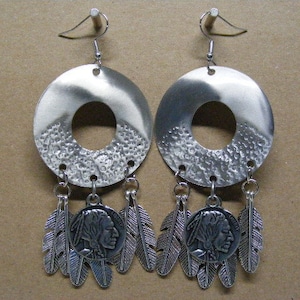 May include: Silver-toned earrings with a circular design and a central hole. The earrings feature a textured lower section, dangling feathers, and a pendant with a Native American profile. The earrings are suspended from a hook.