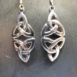 May include: A pair of silver-toned Celtic knot earrings. Each earring features an intricate, interwoven design with a teardrop shape. The earrings hang from a simple hook. The background is a dark, textured surface.