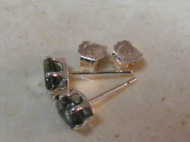 K&ouml;nnte beinhalten: Ein Paar silberne Ohrringe mit gr&uuml;nen Edelsteinen. Die Ohrringe sind im Stecker-Stil mit einem Stift und einem Schmetterlingsverschluss.