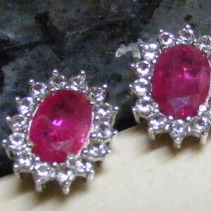 May include: A pair of stud earrings featuring oval-cut, deep pink gemstones surrounded by a halo of smaller, clear stones. The earrings are set in a silver-colored metal, creating a sparkling, elegant design. The earrings are displayed on a light-colored surface.