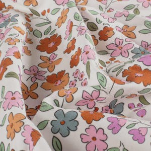 Puede incluir: Primer plano de una tela con estampado floral. La tela es blanquecina con un diseño repetido de flores en tonos naranja, rosa y azul, con hojas verdes. Las flores son estilizadas y están dispersas por el material.
