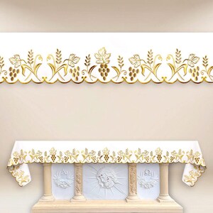 May include: White altar cloth featuring a gold embroidered grape and wheat design. The cloth is draped over a white altar with carved columns and religious symbols. A matching gold embroidered border is at the top.