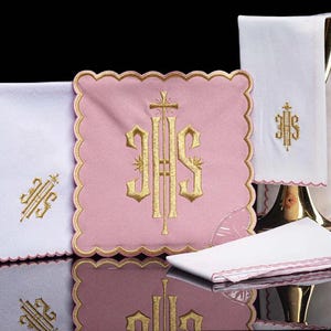 Può includere: Una collezione di tessuti religiosi, tra cui un panno quadrato rosa con ricamo dorato e bordo smerlato. Sono presenti anche panni bianchi con ricamo dorato e bordi smerlati, insieme a un panno bianco piegato con un bordo rosa.