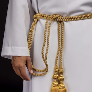 Peut inclure: Une ceinture en corde dorée avec des glands, nouée autour d'un vêtement blanc. La ceinture est faite de corde torsadée et comporte des glands décoratifs aux extrémités. L'image est un gros plan de la ceinture et du vêtement.