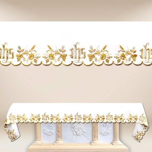 May include: A white altar cloth with gold embroidered floral and IHS designs. The cloth drapes over a white altar with carved religious symbols. The top of the image features a matching gold embroidered border.