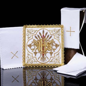 Puede incluir: Ropa de altar religiosa blanca con bordados dorados. La pieza central presenta un diseño ornamentado con las letras IHS, trigo y uvas. Una cruz dorada está bordada en las otras piezas. El conjunto se exhibe sobre una superficie reflectante.