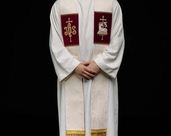 May include: A white religious garment with a cream-colored stole featuring burgundy panels with gold embroidery. The stole has gold fringe at the bottom. The person is wearing a white turtleneck and black shoes.